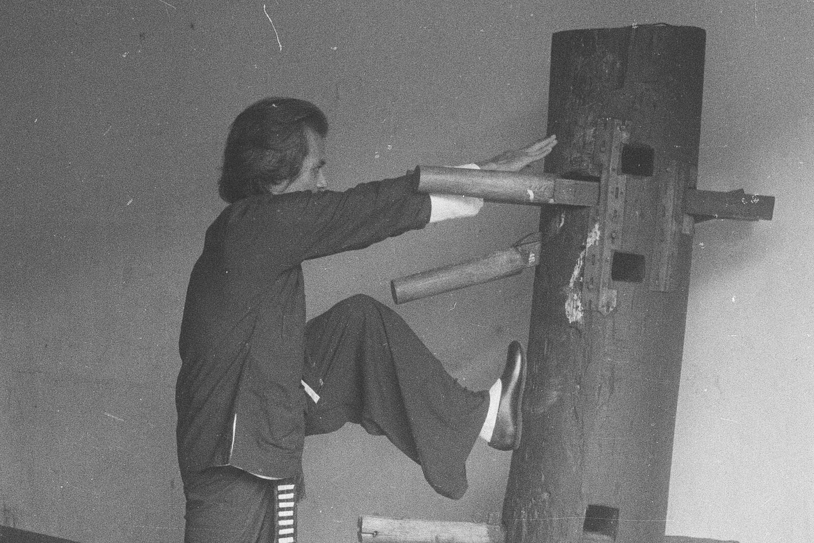 Photographie en noir et blanc : exercice au mannequin de bois, bras tendus et jambe levée